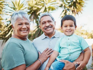 Smiling grandparents holding their young grandson outdoors on a sunny day.