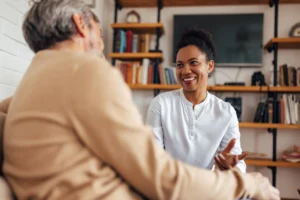 Friendly caregiver smiling and talking with an elderly man at home, providing companionship and emotional support.